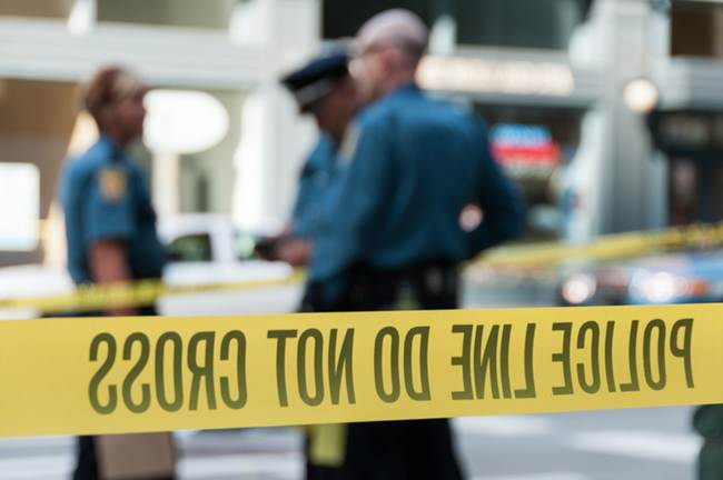 Police stand behind yellow tape at a crime scene. Text on the tape reads “POLICE LINE DO NOT CROSS”.